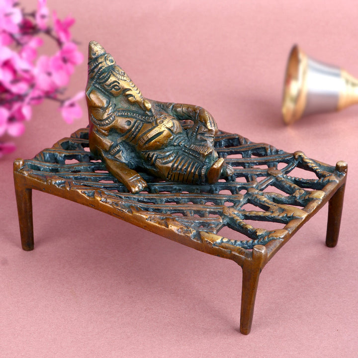 Antique brass Ganesha idol depicting Lord Ganesha relaxing on a traditional charpoy, intricately detailed to symbolize tranquility, wisdom, and divine blessings. Perfect for spiritual décor or worship. India’s Largest Pooja Accessories Brand – www.satvikworld.com